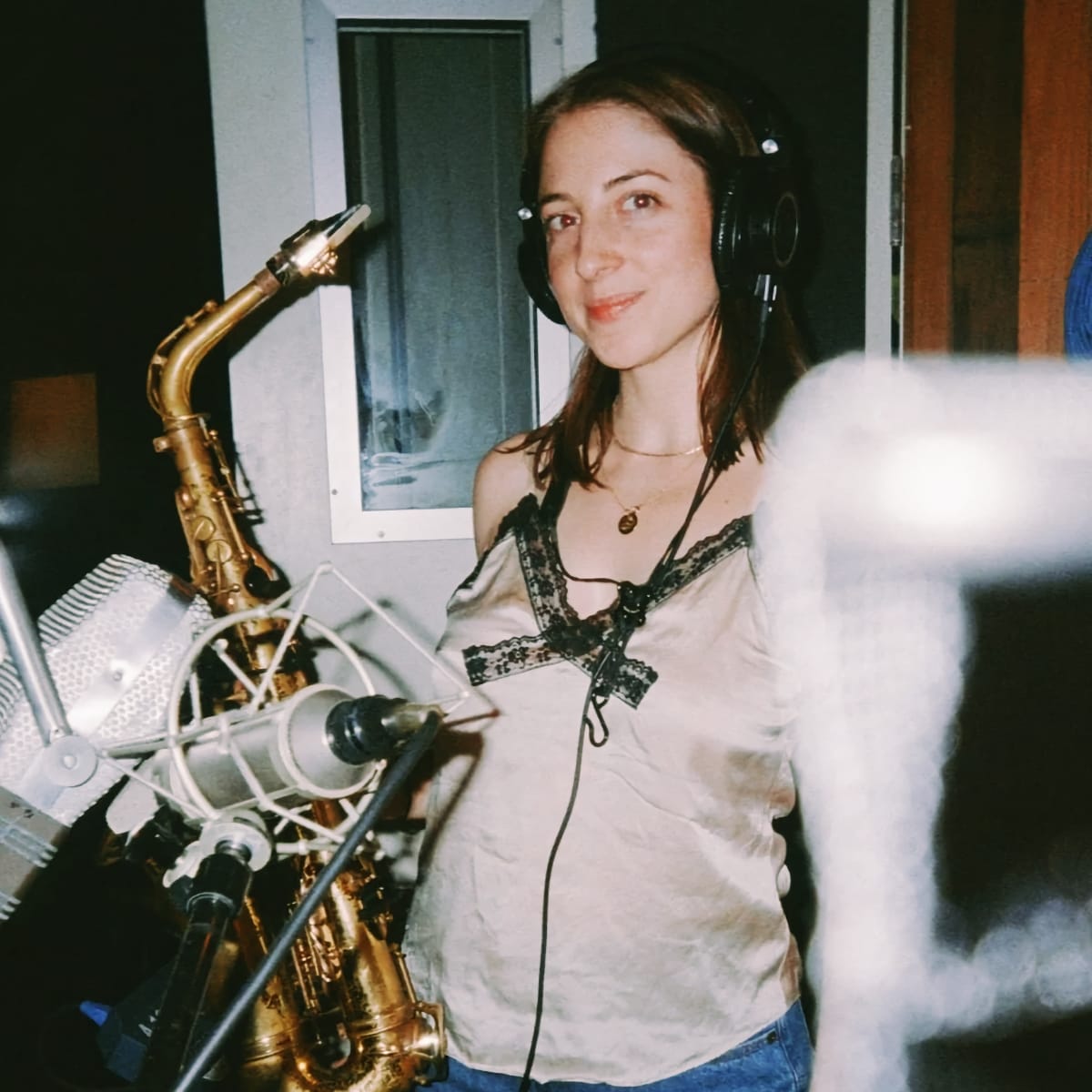 Alden Hellmuth wears headphones and stands beside a saxophone on a mic stand in a recording studio, smiling toward the camera.