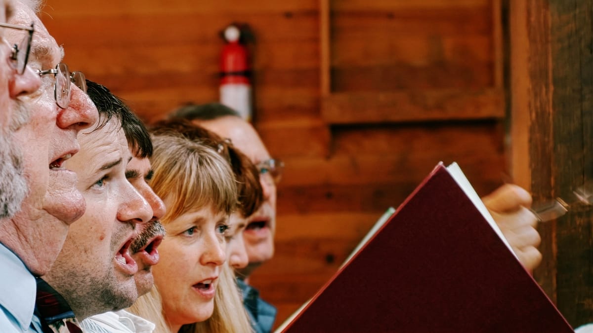 Singers on the front bench of the tenor section at the Holly Springs Sacred Harp convention, mouths open mid-song, hymnals raised, in a wood-paneled hall.
