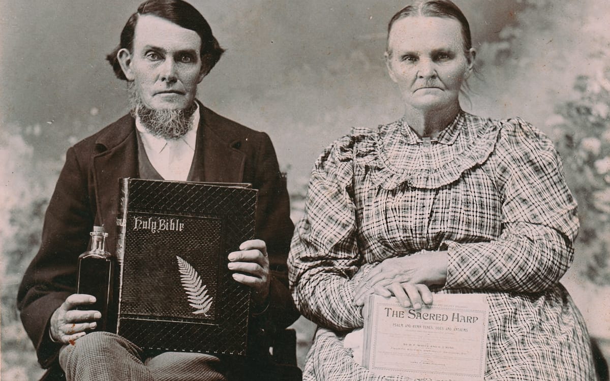 Isaac and Mary McCendon pose for a formal portrait, he holding a Holy Bible and a small bottle, she with a copy of The Sacred Harp on her lap. Sepia-toned, 1800s.