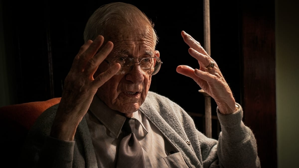 Raymond Hamrick speaks mid-sentence, both hands raised, set against a dark background.