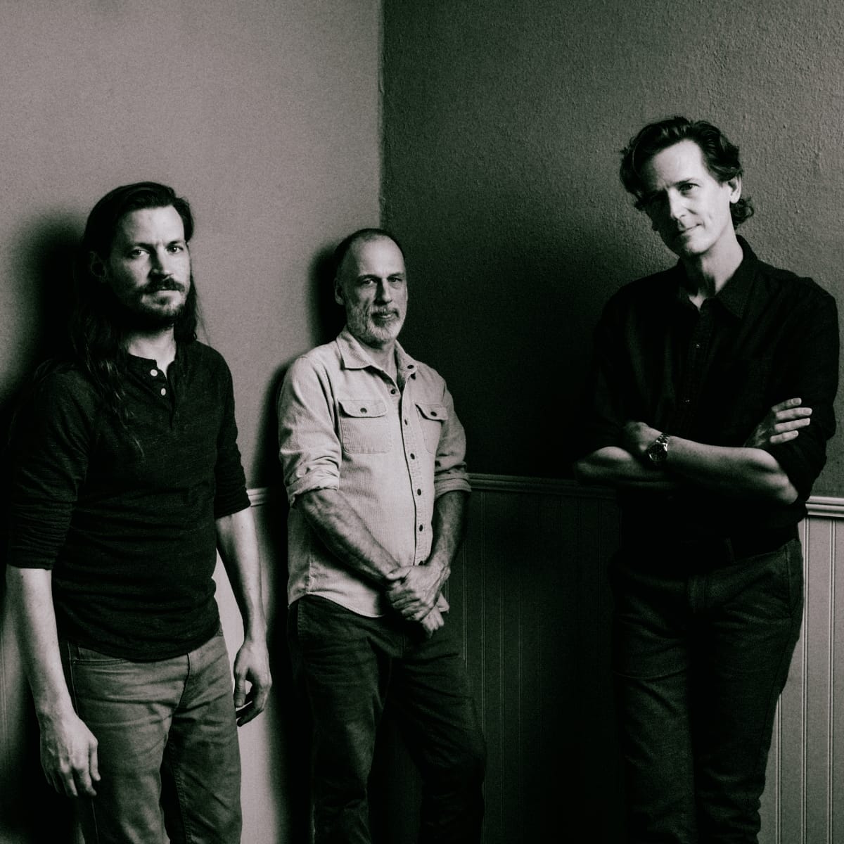 Cento Threeo in a black-and-white portrait, three members standing against a corner wall, one with arms crossed at right, one centered with hands clasped, one at left looking aside.