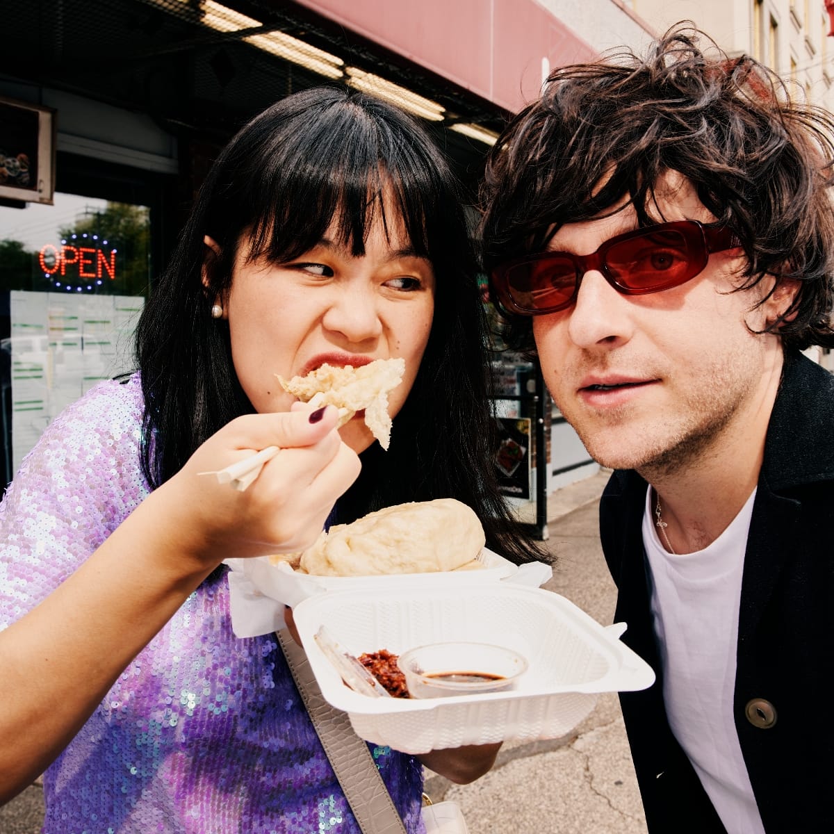 Anne Tong eats a dumpling with chopsticks outside a restaurant as Bryce Barsten looks on, both of Chinese American Bear, Tong in a lavender sequined top, Barsten in red sunglasses.