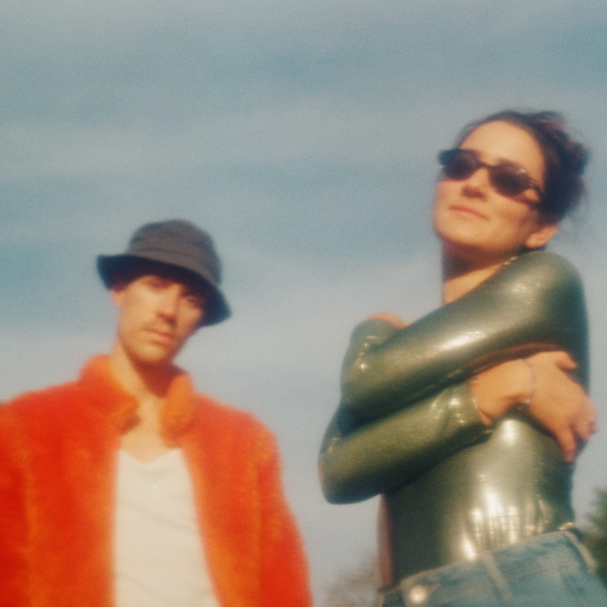 Curió Curió outdoors against a pale sky; one figure in an orange jacket and dark hat stands blurred behind, the other in sunglasses and a glossy olive top facing the camera.