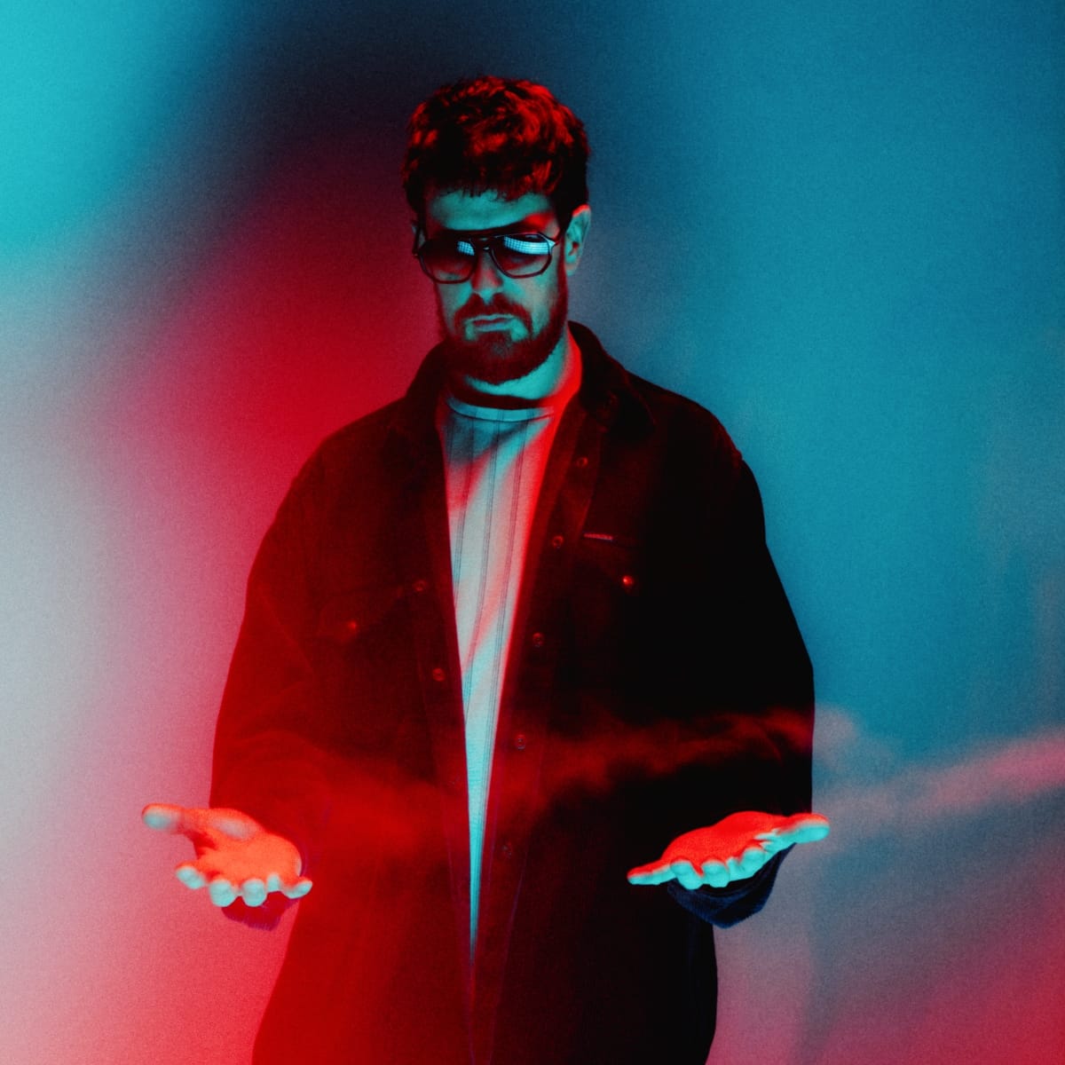 Danalogue stands with both palms open and outstretched, wearing dark LED-lit glasses and an open jacket, lit in red and teal against a smoky gradient background.
