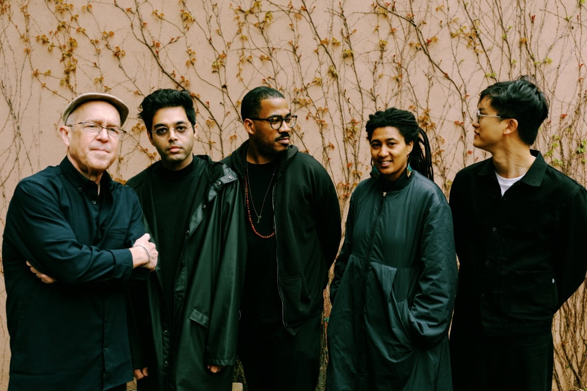 Dave Douglas, Rafiq Bhatia, James Brandon Lewis, Tomeka Reid, and Ian Chang stand against a pale wall threaded with bare vines, all in dark clothing, gazing in different directions.