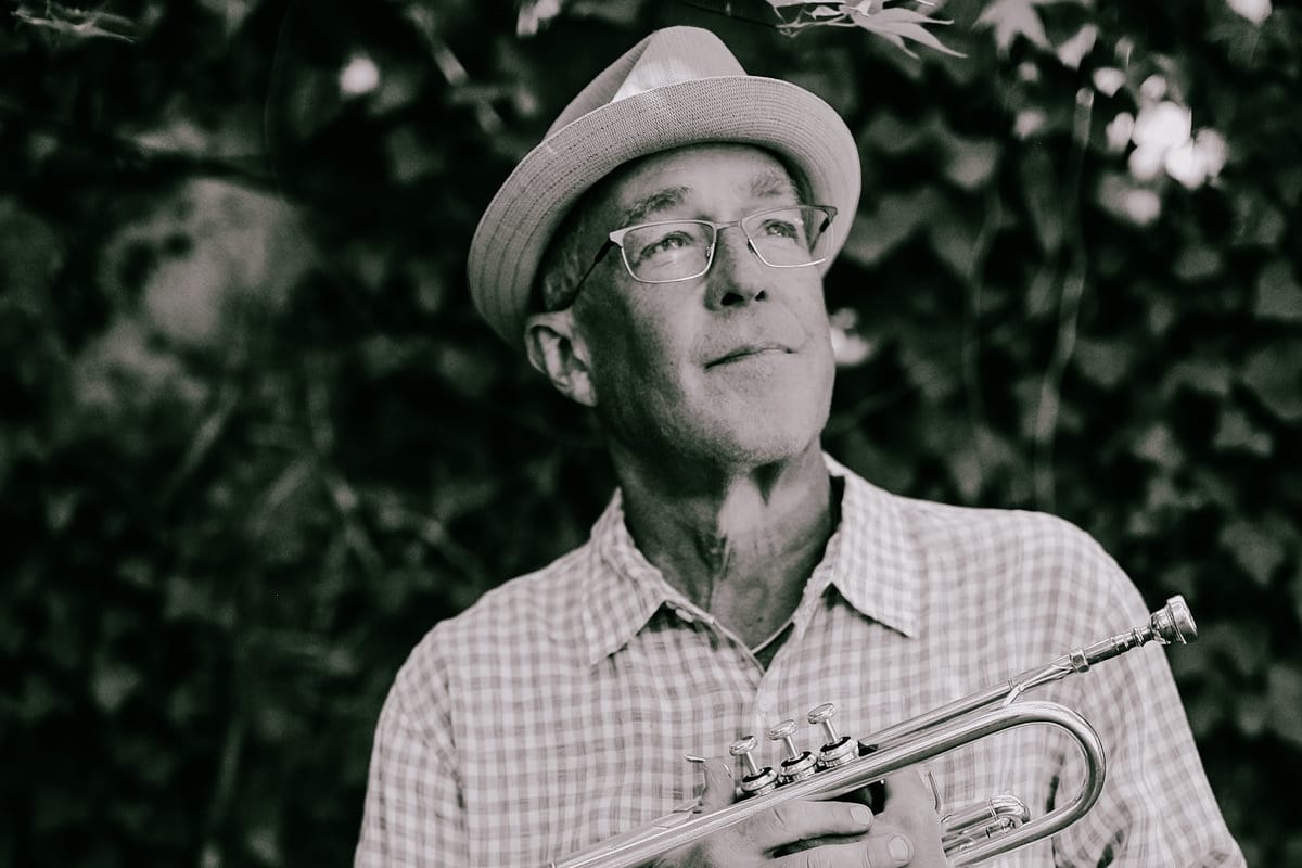 Dave Douglas holds a trumpet at his side, looking upward, wearing a straw hat and checked shirt, with ivy-covered foliage blurred behind him.