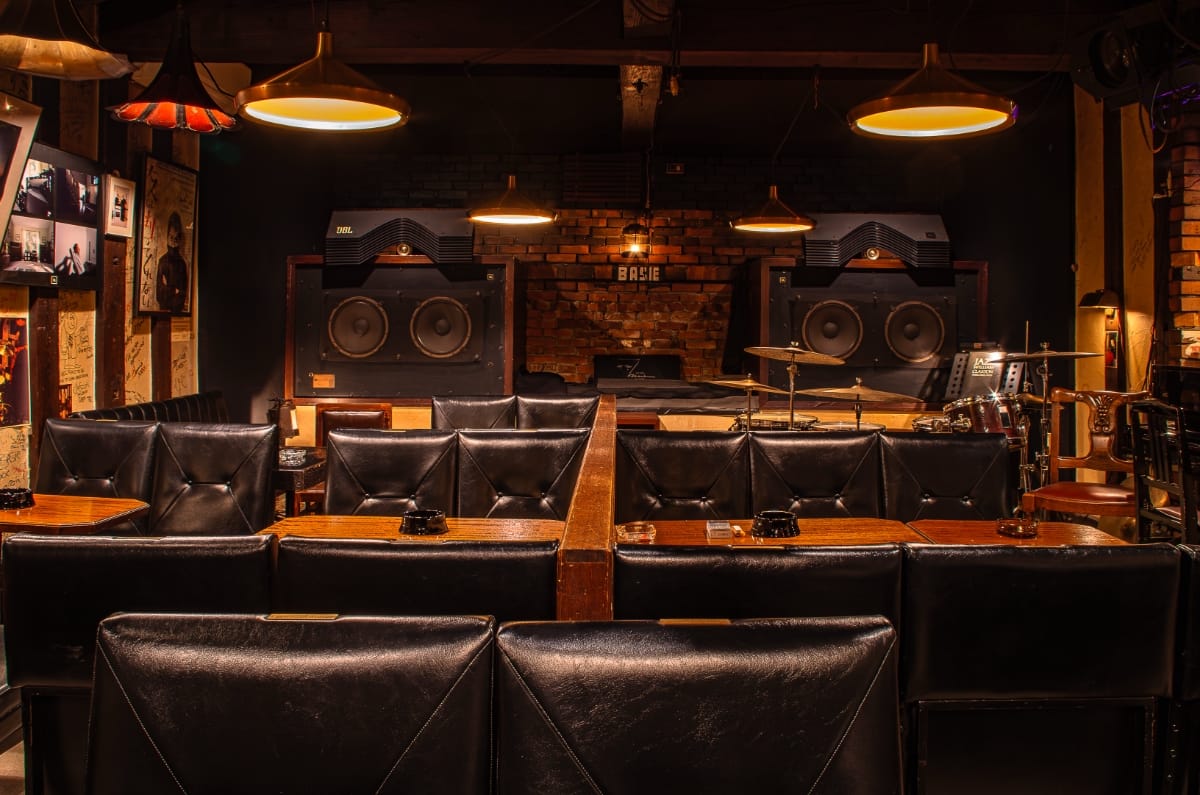Rows of black tufted leather booths face two massive JBL speakers mounted on an exposed brick wall inside jazz kissa Basie, copper pendant lamps overhead, a drum kit visible to one side.