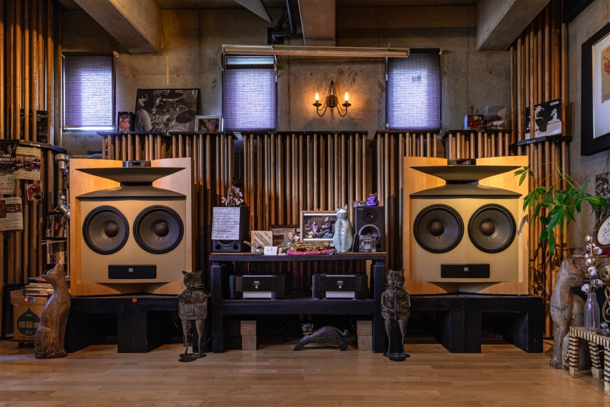 Two large JBL speakers in maple cabinets dominate jazz kissa Candy's listening room, flanked by bronze cat figurines — and a real cat stretched flat under the equipment stand between them.