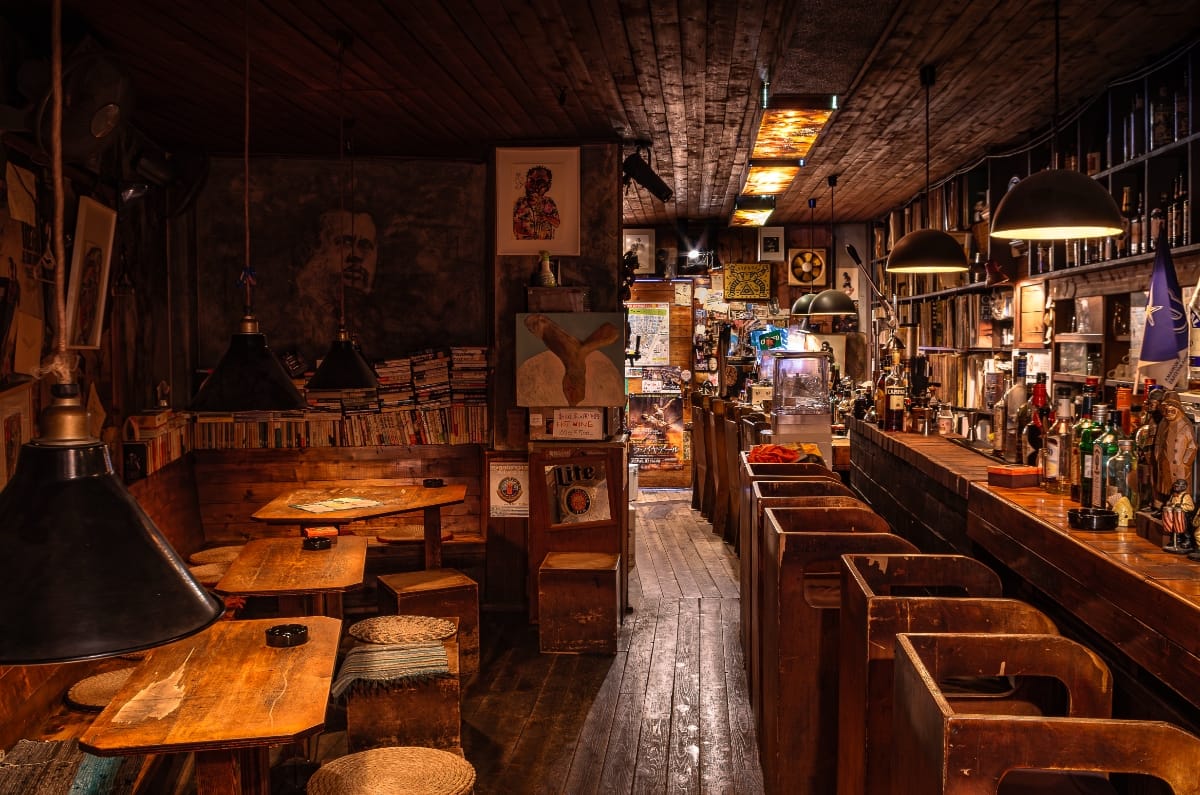Jazz kissa Minton House: a narrow room with wooden tables and stools on the left, a long bar lined with bottles on the right, walls dense with jazz posters and artwork, black pendant lamps overhead.