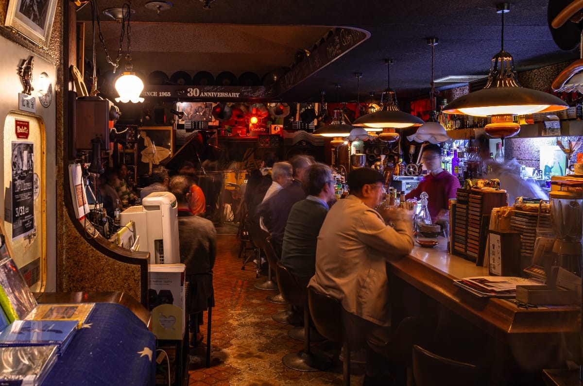 A busy evening at jazz kissa Roulette: customers packed along a long bar and spilling into the room, a bartender working behind the counter, vinyl records lining the ceiling's edge overhead.