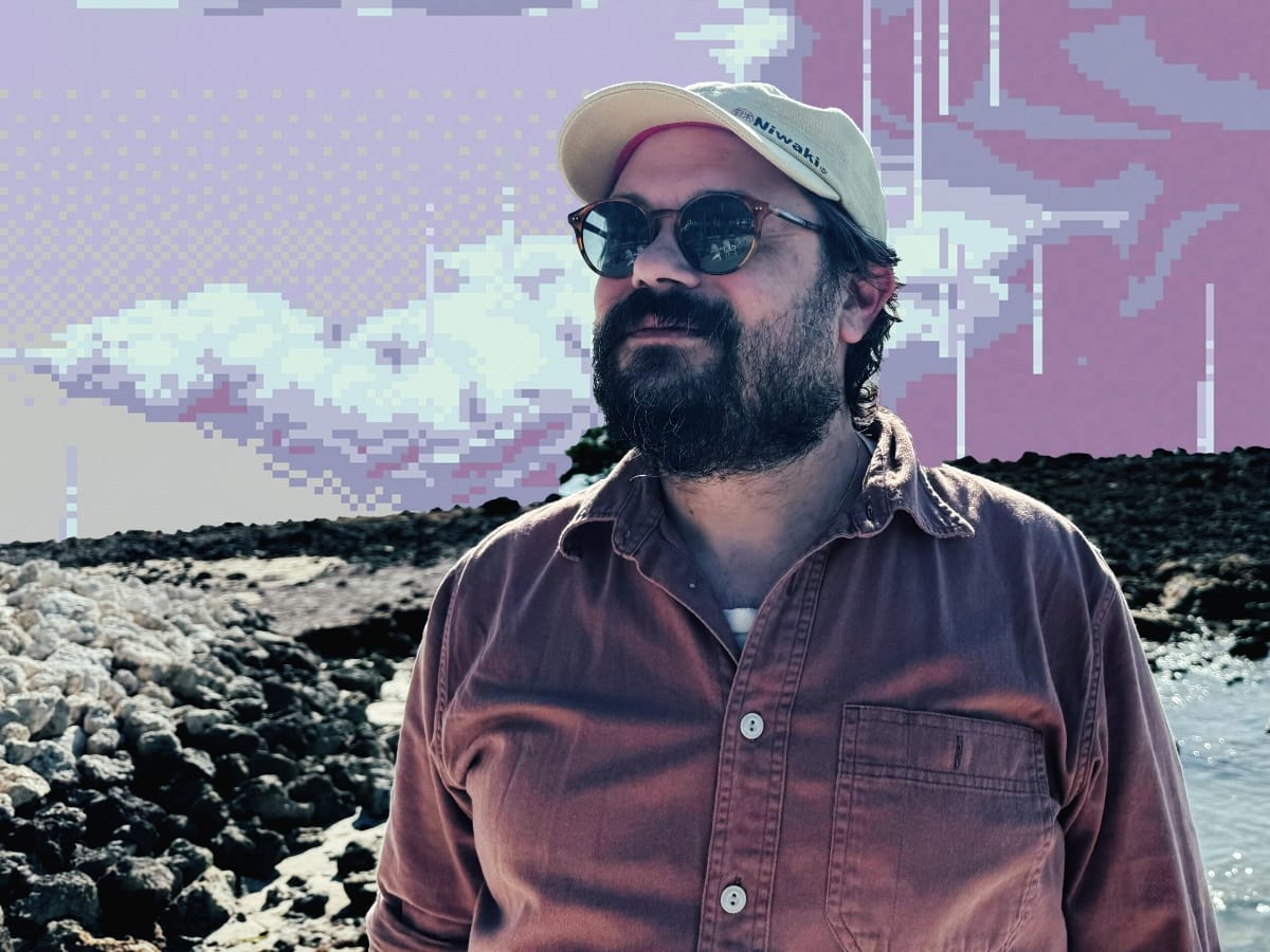 M. Geddes Gengras stands at a rocky shoreline in a cap and round sunglasses against a pixelated, glitch-art sky of lavender and pink clouds.