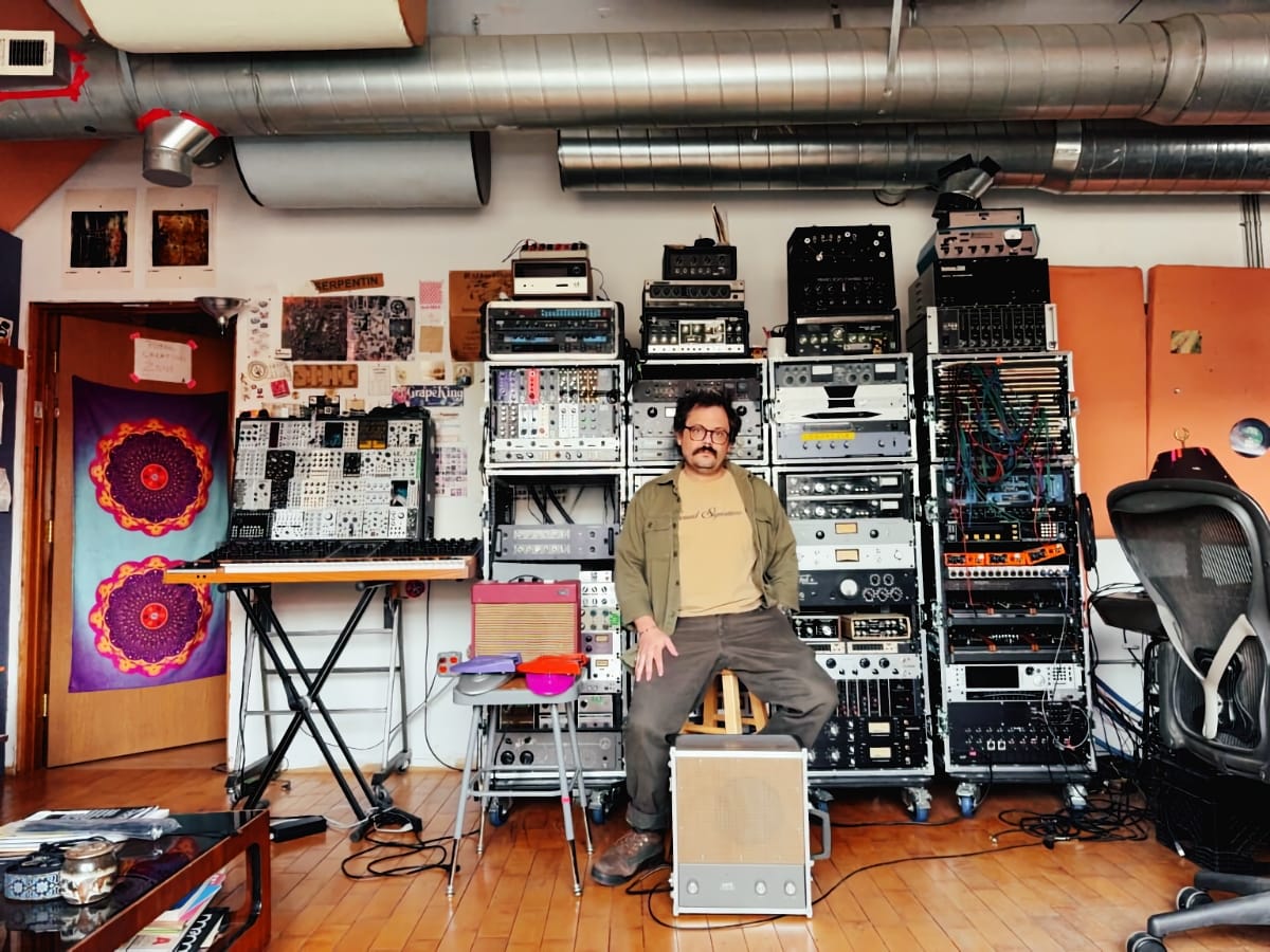 M. Geddes Gengras sits on a stool in his studio, surrounded by floor-to-ceiling racks of synthesizers and recording gear, a modular synth rig to his left.