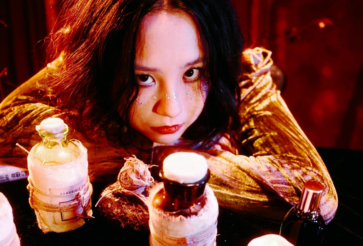 Mixol rests her arms on a dark table of gauze-wrapped apothecary bottles, gazing at the camera in a shimmering gold top with glitter beneath her eyes, in warm red light.