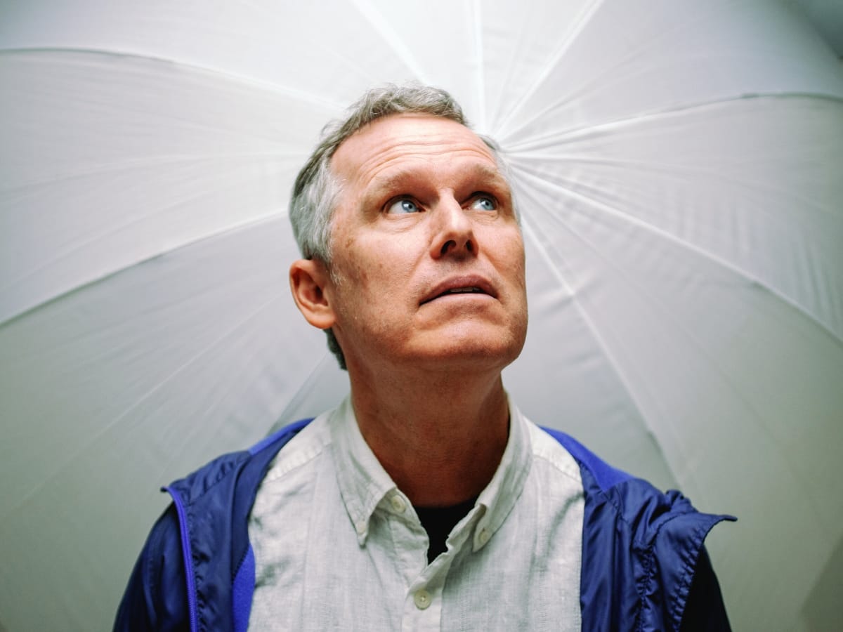 Matthew S of Myth Math looks upward beneath a large white umbrella that fills the frame behind him, wearing a blue windbreaker over a pale button-up shirt.