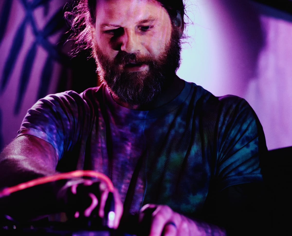 Nolan Hildebrand, bearded and in a tie-dye shirt, leans over a table of cables and knobs, both hands on the controls, lit in magenta and teal.