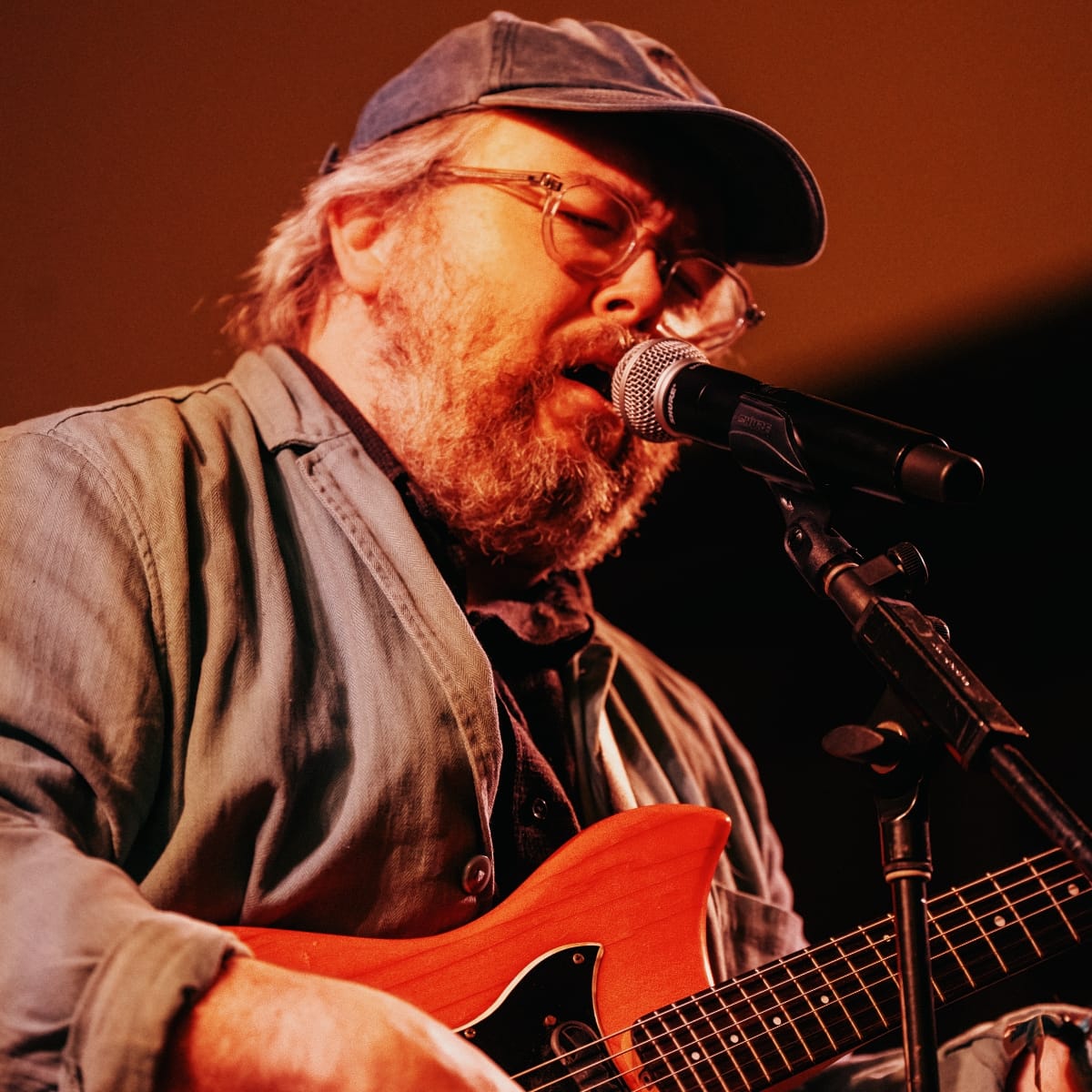 Richard Dawson sings into a microphone while playing a red electric guitar, eyes closed, warm stage light on his beard and cap.