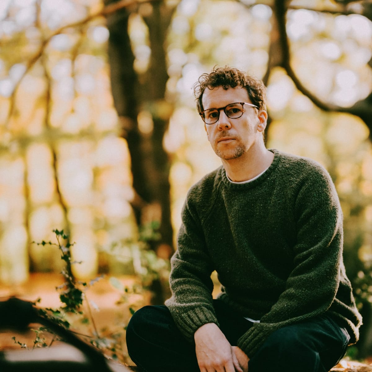 Scrimshire seated on a log in a sunlit woodland, wearing glasses and a dark green knit sweater, looking directly into the camera.
