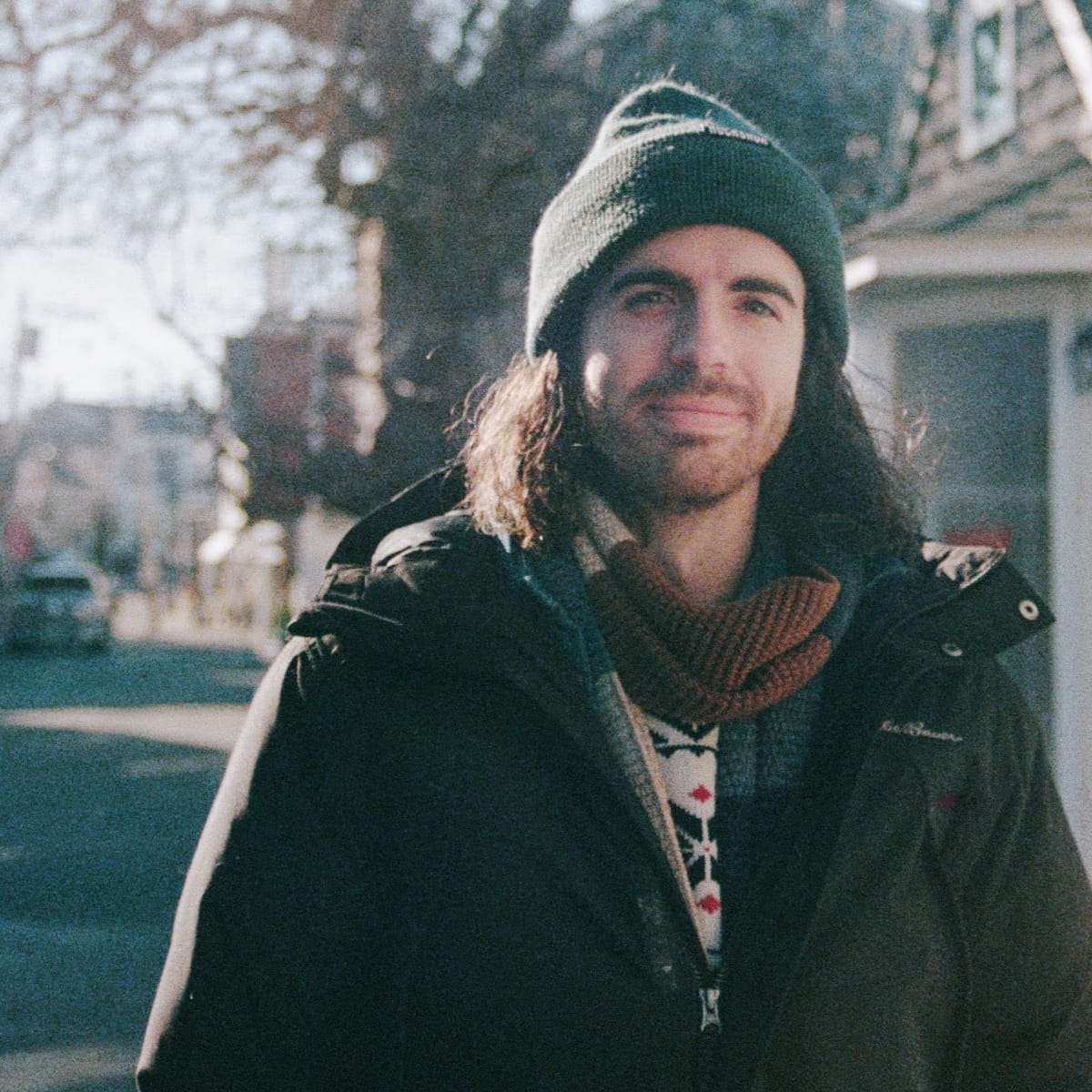 St. Silva stands on a sunlit street in a green knit beanie, dark parka, and patterned sweater, half-smiling toward the camera.
