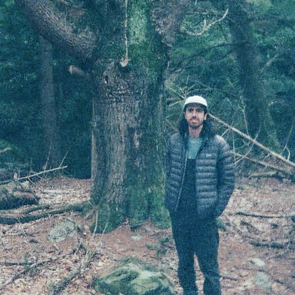 St. Silva stands before a towering moss-covered tree in a bare winter forest, hands tucked into a dark puffer jacket, a white cap on his head.