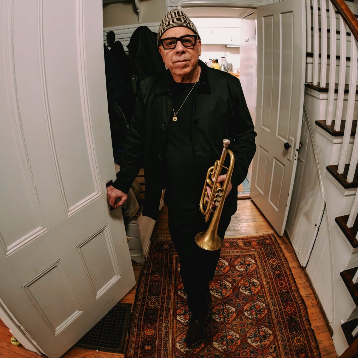 Steven Bernstein stands in a doorway holding a trumpet at his side, wearing a black jacket, glasses, and a patterned knit cap, a Persian rug and staircase behind him.
