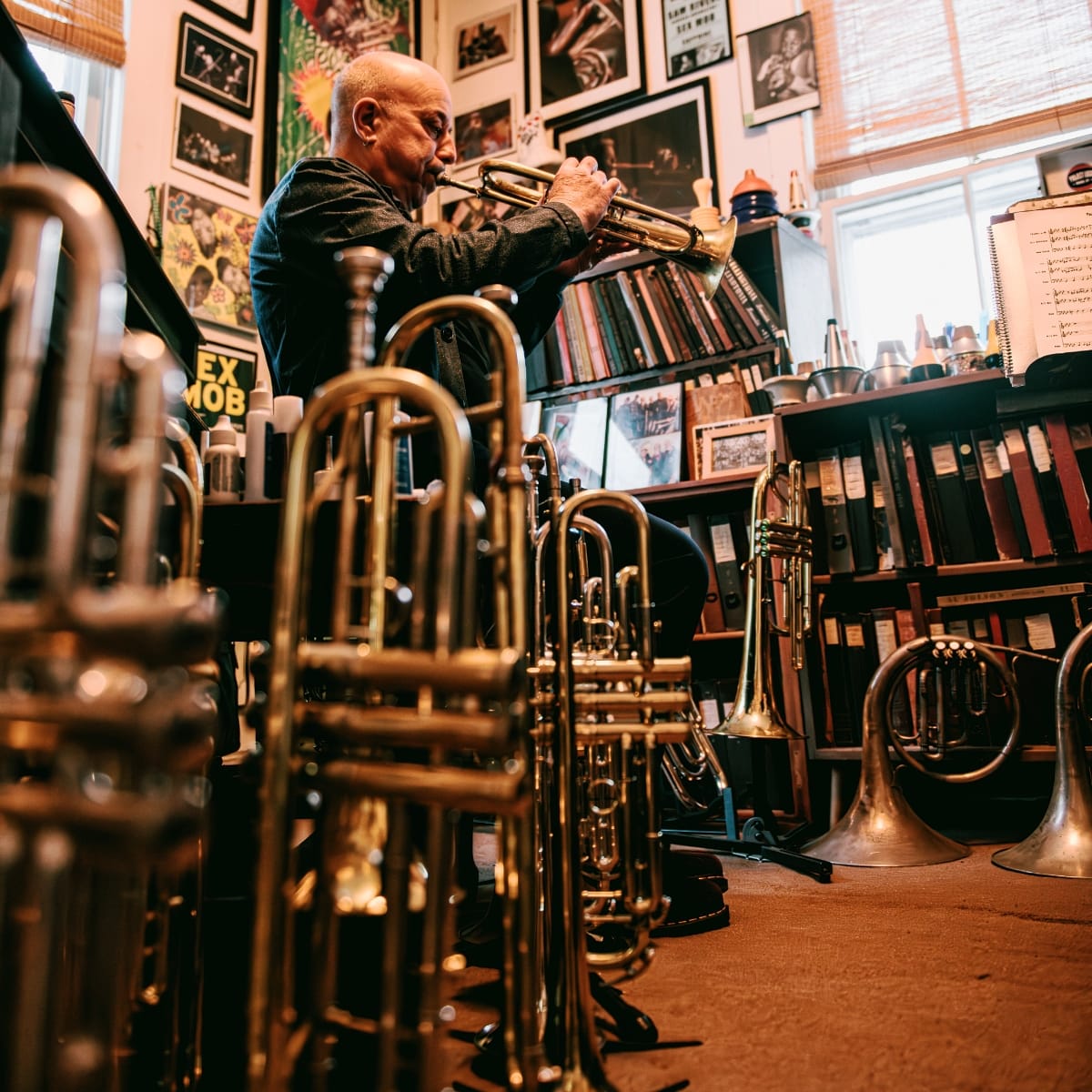 Steven Bernstein plays trumpet in profile in a studio crowded with brass instruments in the foreground, framed photos and record posters covering the walls, and shelves of books and binders behind him.