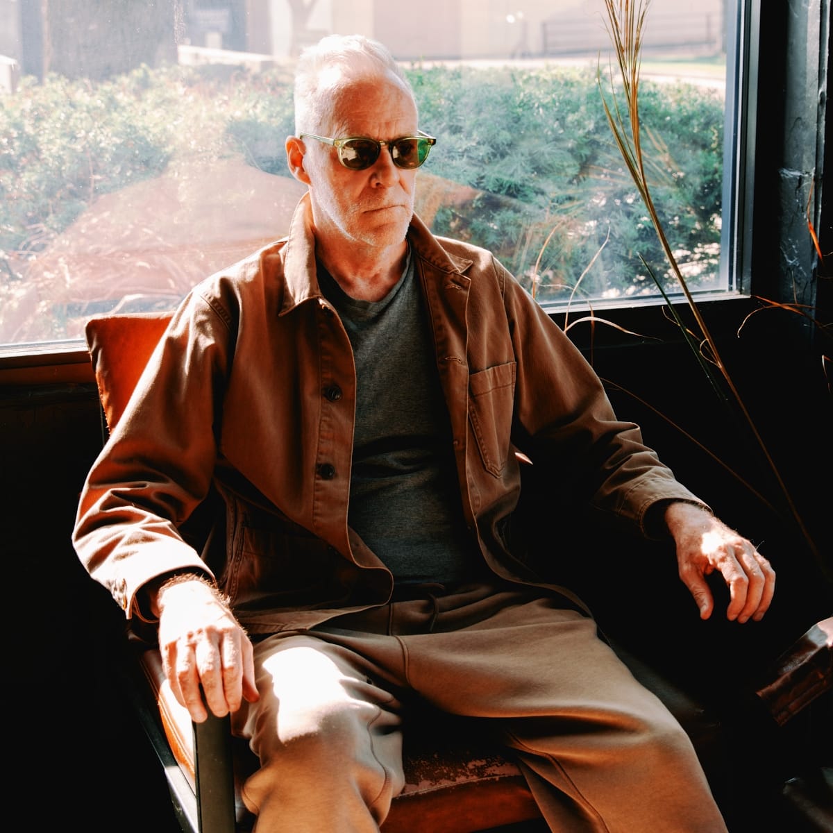 Steven Hall sits in a wooden armchair by a large window in sunglasses and a brown jacket over a grey T-shirt, one hand resting on the armrest, greenery and sunlight behind him.