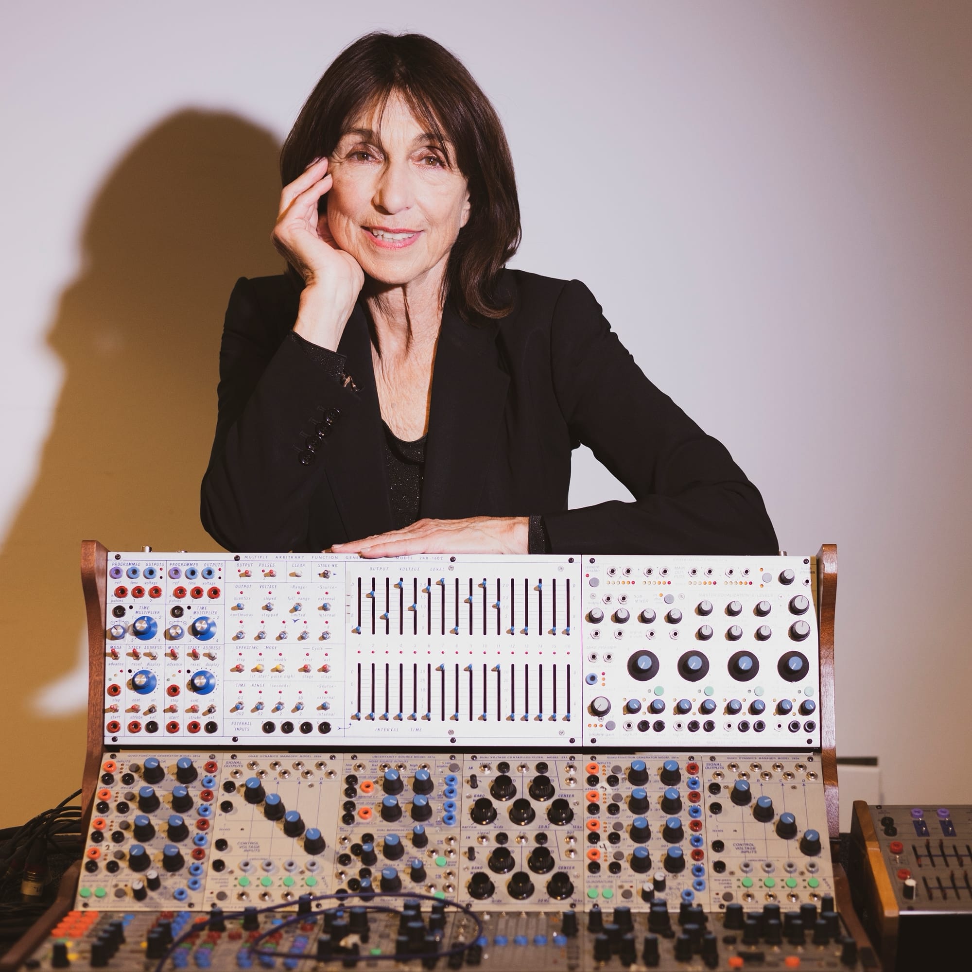Suzanne Ciani rests her chin on one hand and smiles at the camera, seated behind a large wood-framed Buchla modular synthesizer covered in knobs, sliders, and colored patch jacks, her shadow on the wall behind.