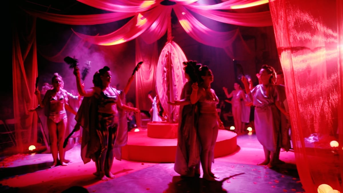 Bathed in red light, The Goddess Party performers in draped grey garments hold feathers around a raised platform topped with a glowing oval sculpture, sheer red curtains overhead.
