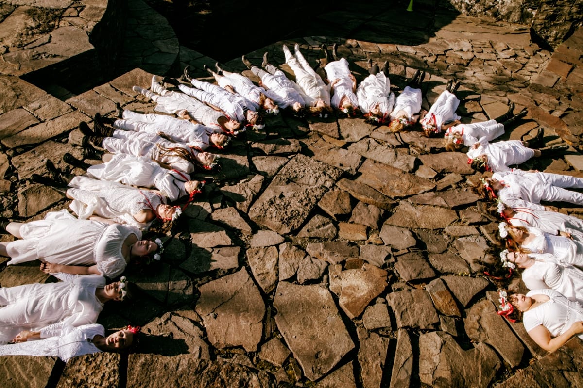 The Goddess Party performers dressed in white with flower crowns and red ribbons lie in a radiating circle on sunlit flagstone, heads toward the center, seen from above.