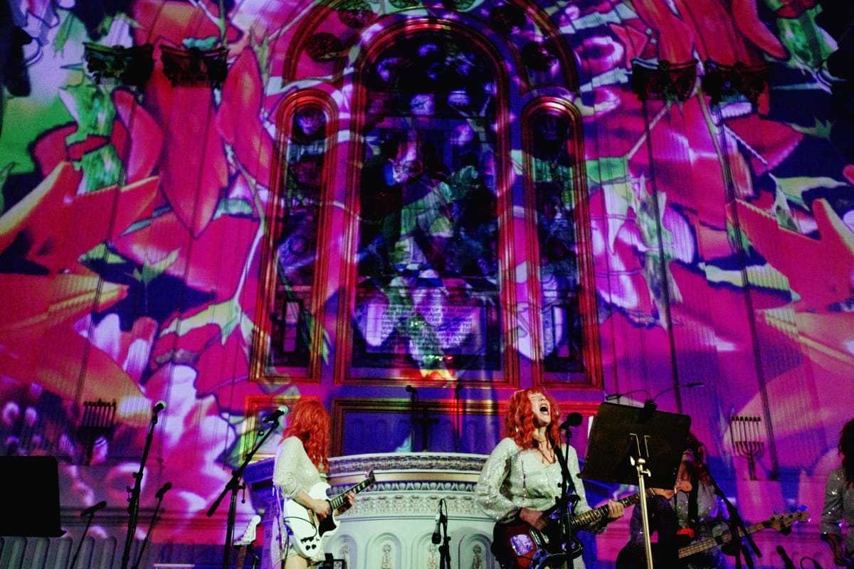 Under giant projected pink lilies on a stained-glass church wall, a red-haired singer in sequins belts into a microphone while playing electric guitar, flanked by two guitarists.