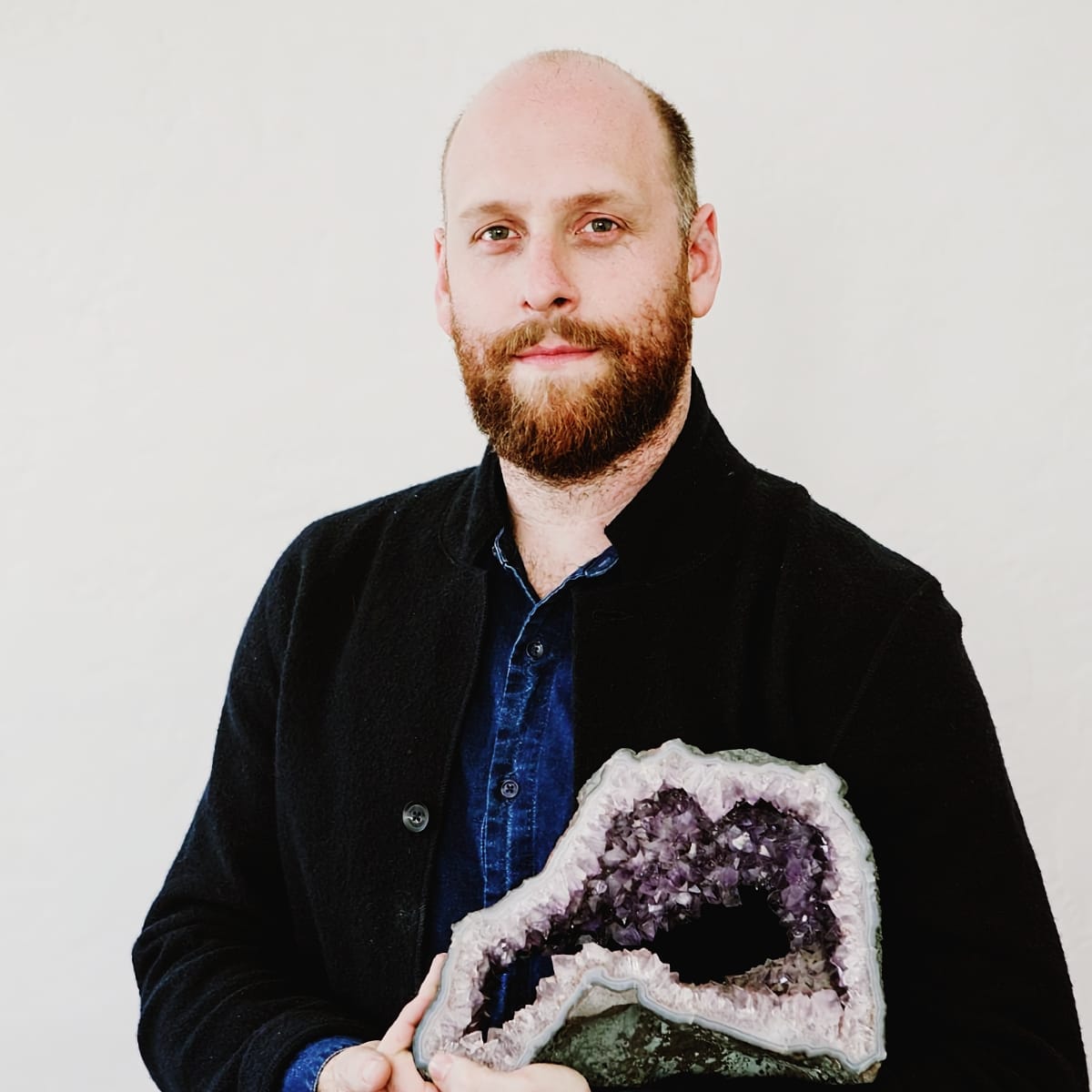 Chris Rountree holds a large amethyst geode against a plain white background, wearing a black jacket over a blue shirt.
