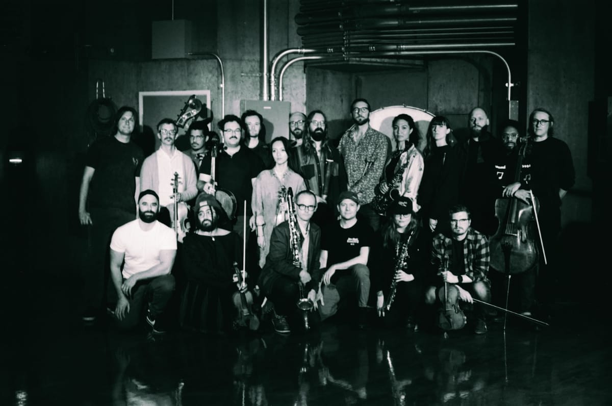 Wild Up ensemble, roughly twenty musicians posed in two rows in a dimly lit industrial space, many holding instruments including strings, saxophones, and brass, shot in black and white.