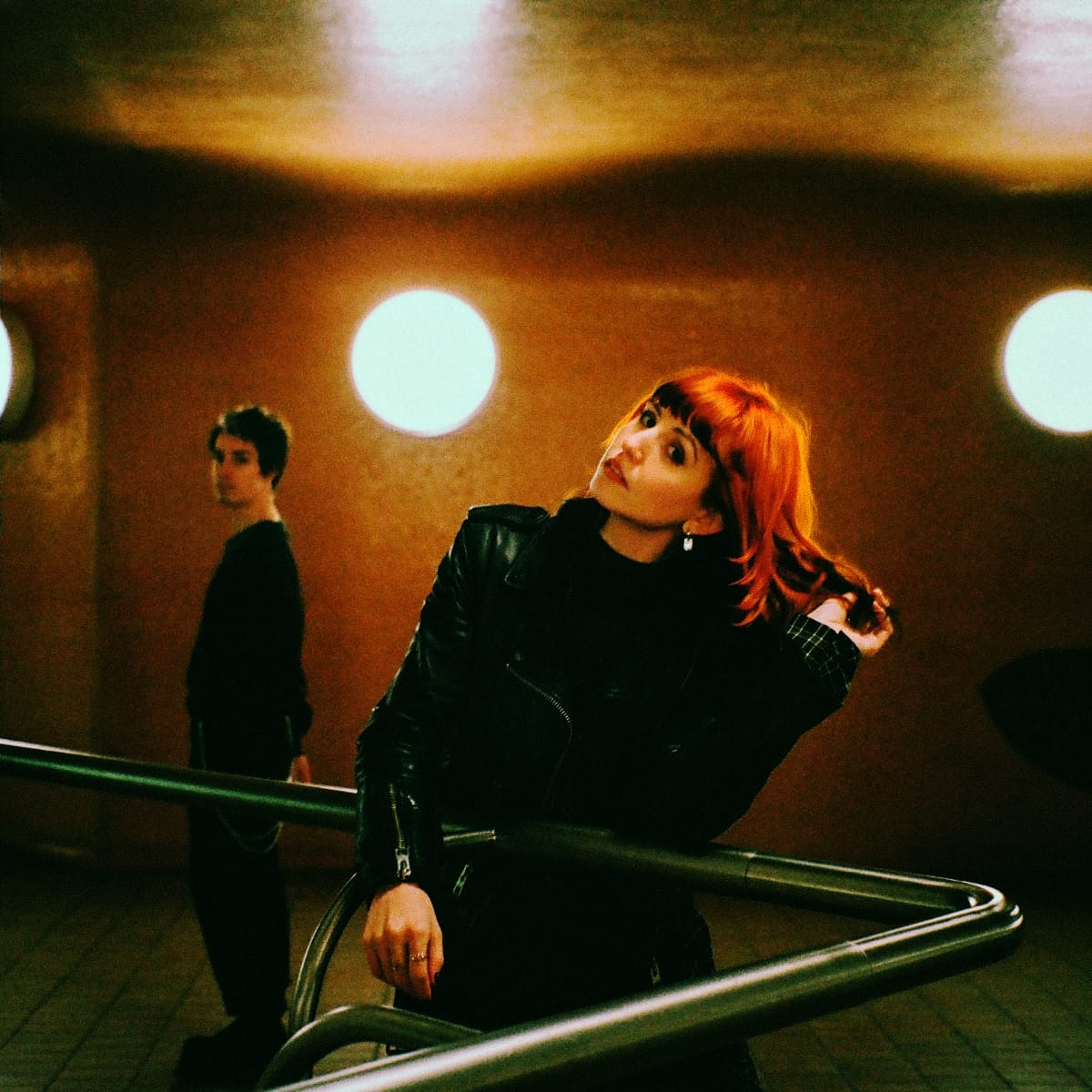 Courtney Carmichael of sundayclub leans over a metal railing under warm round lights, red hair catching the glow, with Nikki St. Pierre standing apart in the background.