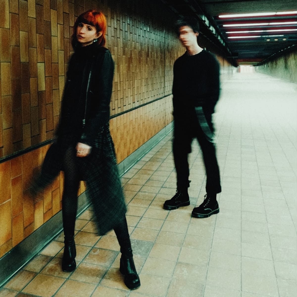 Courtney Carmichael and Nikki St. Pierre of sundayclub stand in a tiled underground passage, Carmichael in focus with red hair, St. Pierre blurred with motion behind him.