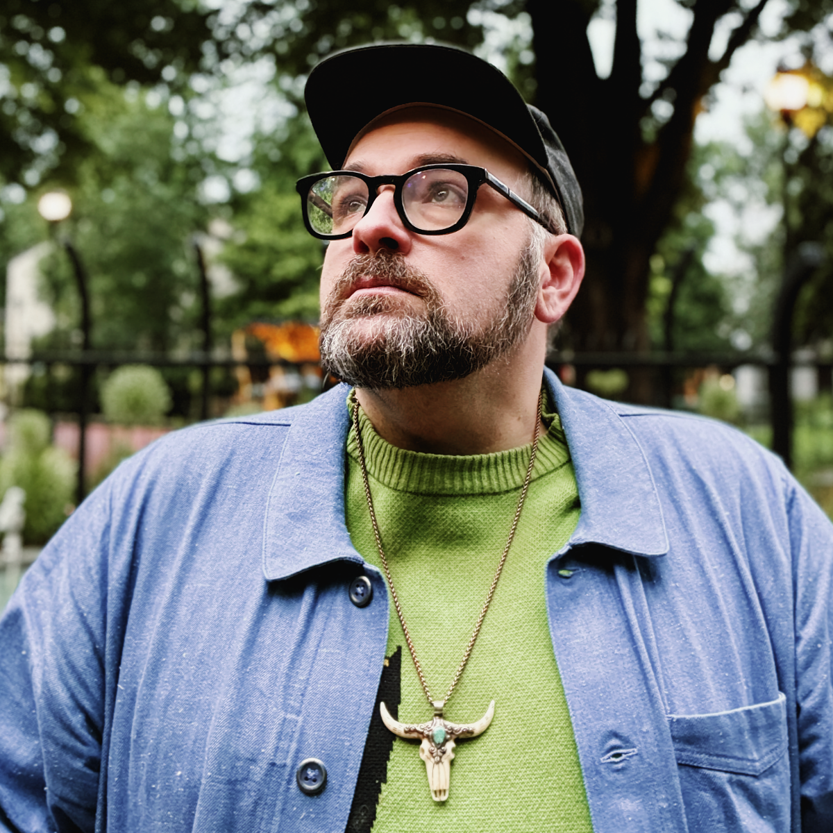 Tyler Pursel looks off to one side in a park, wearing a backwards cap, dark glasses, a green sweater under a blue chore jacket, and a longhorn skull pendant.