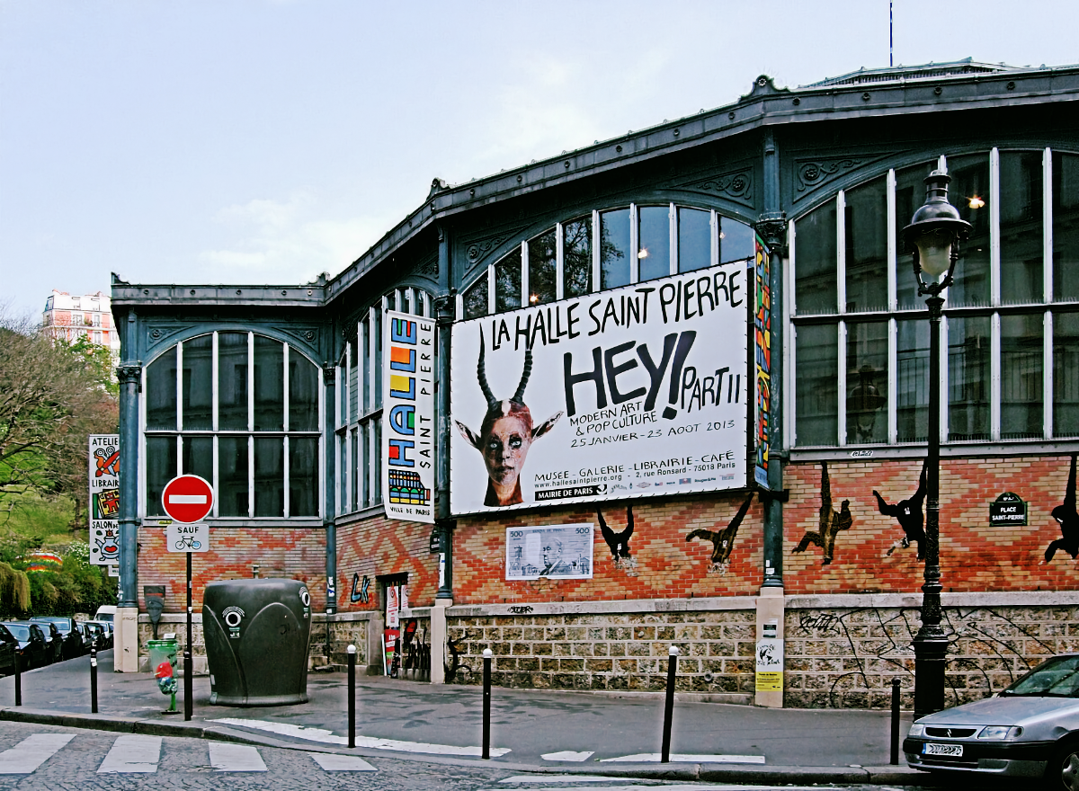 The Halle Saint Pierre in Montmartre, an iron-and-glass market hall in red brick, its facade hung with a banner for the HEY! Part II exhibition showing a face with antelope horns.