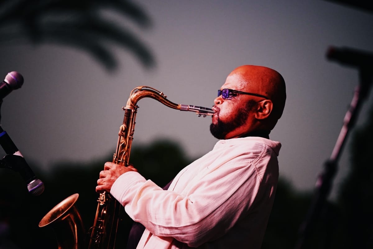 Azar Lawrence plays tenor saxophone in profile onstage at dusk, framed by microphone stands.