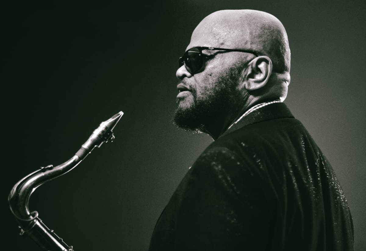 Azar Lawrence in profile against a dark backdrop, sunglasses on, the neck and mouthpiece of his tenor saxophone rising in the foreground.