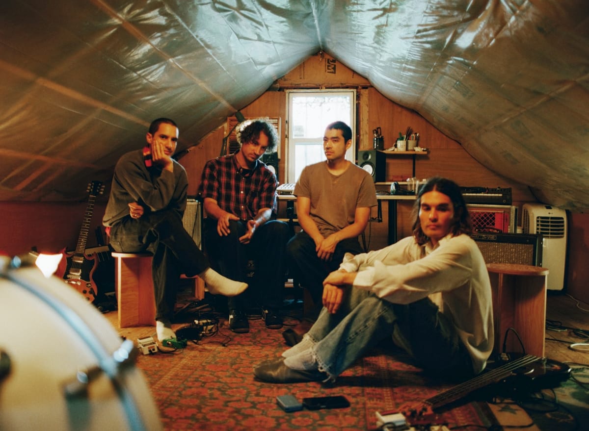 Bondo sit and lean around an attic practice room lined with plastic sheeting, guitars, amps, and pedals scattered across a patterned rug, a drum head filling the foreground.