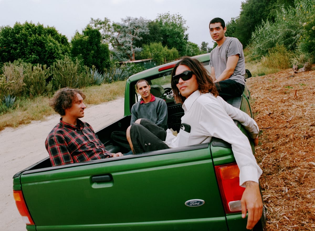 Bondo gather in and around the bed of a green Ford pickup parked on a dirt path, cactus and dry brush rising on the slope behind them.