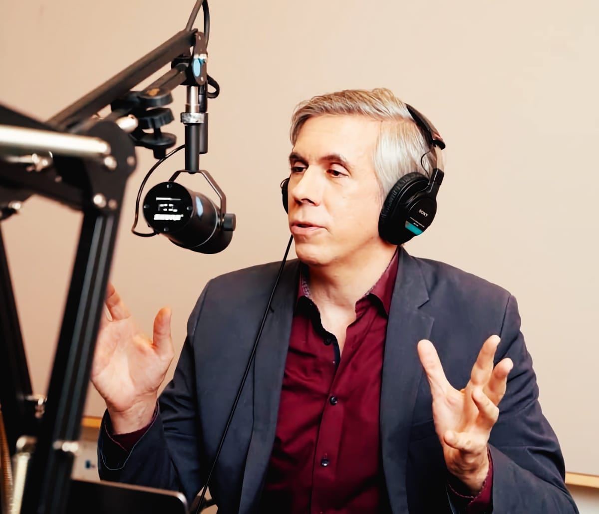 Chris Molanphy speaks into a studio microphone wearing headphones and a navy blazer over a burgundy shirt, both hands raised mid-gesture.