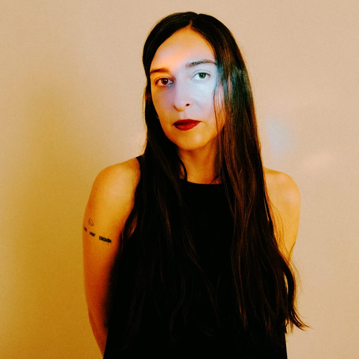 Concepción Huerta faces the camera against a warm tan wall, long dark hair loose over a black sleeveless top, a cool blue light falling across one side of her face.