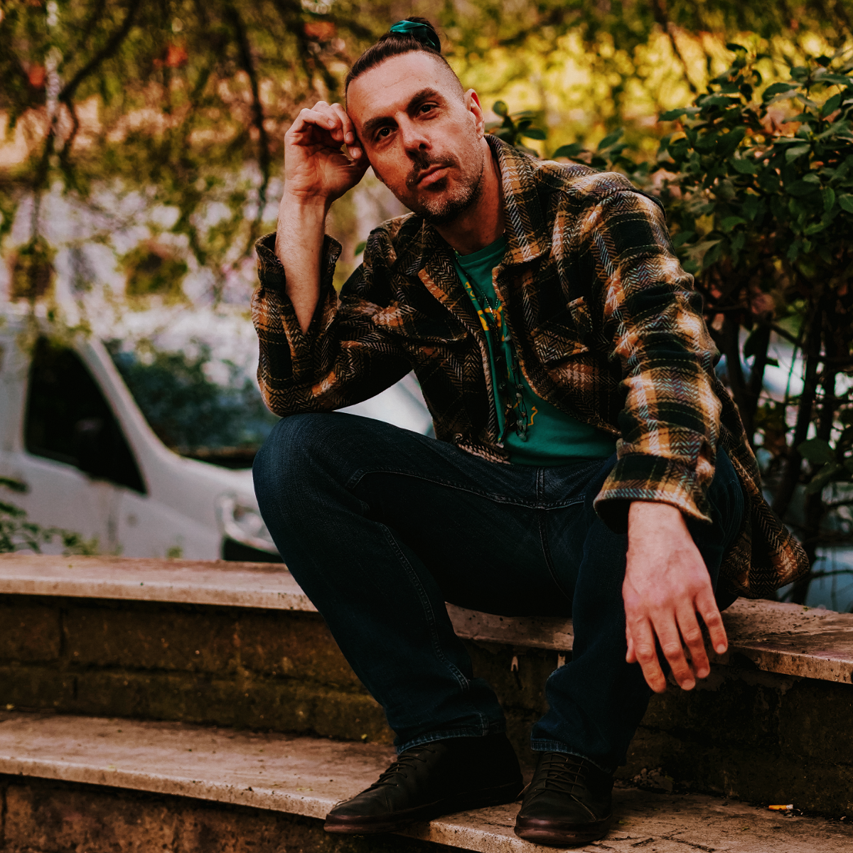 Dave De Rose sits on a low stone step in a plaid wool jacket and jeans, one hand at his temple, looking directly at the camera. Green foliage and a parked white van blur behind him.