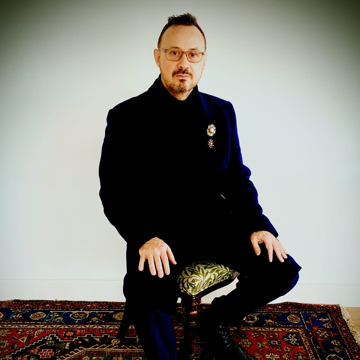 David Norland sits on a small leaf-patterned stool on a red Persian rug, hands on his knees, wearing a dark coat with two ornate jeweled brooches at the lapel.