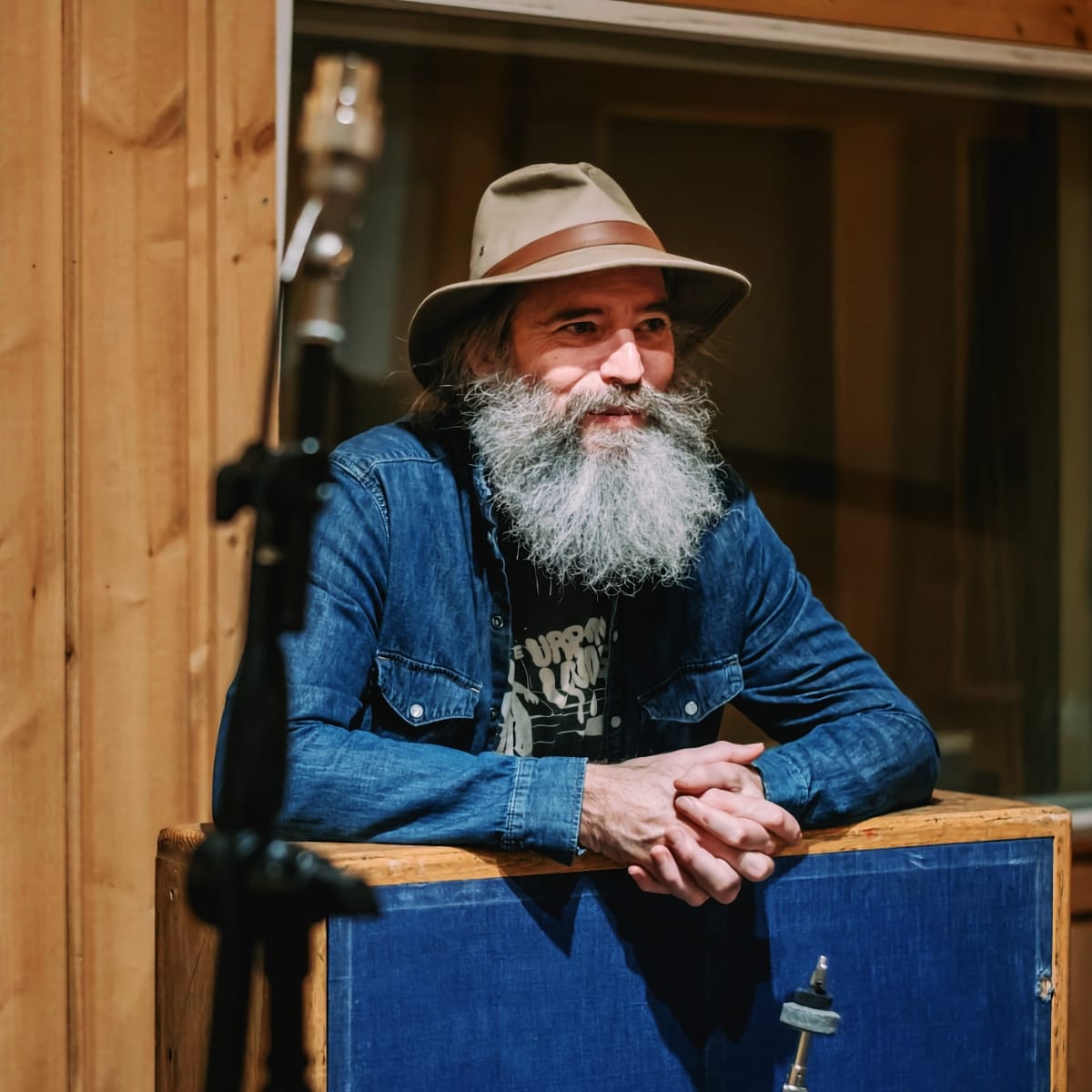 Ethan Miller leans on a padded studio baffle, hands clasped, looking off to one side. Wide-brimmed hat, denim shirt, long beard; a mic stand and wood panelling behind him.
