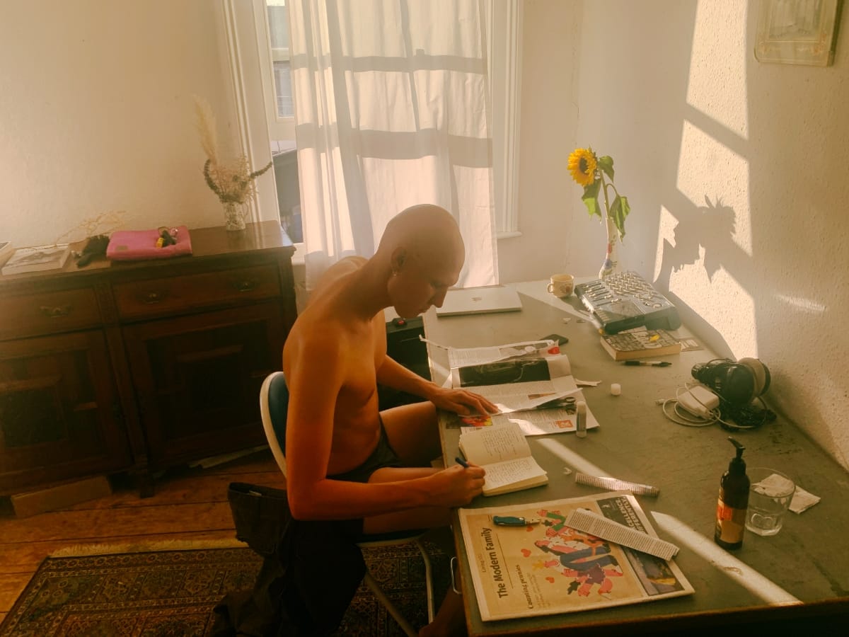 GB sits shirtless at a sunlit desk, writing in an open notebook surrounded by spread newspaper pages and clippings, a sunflower in a vase behind him.