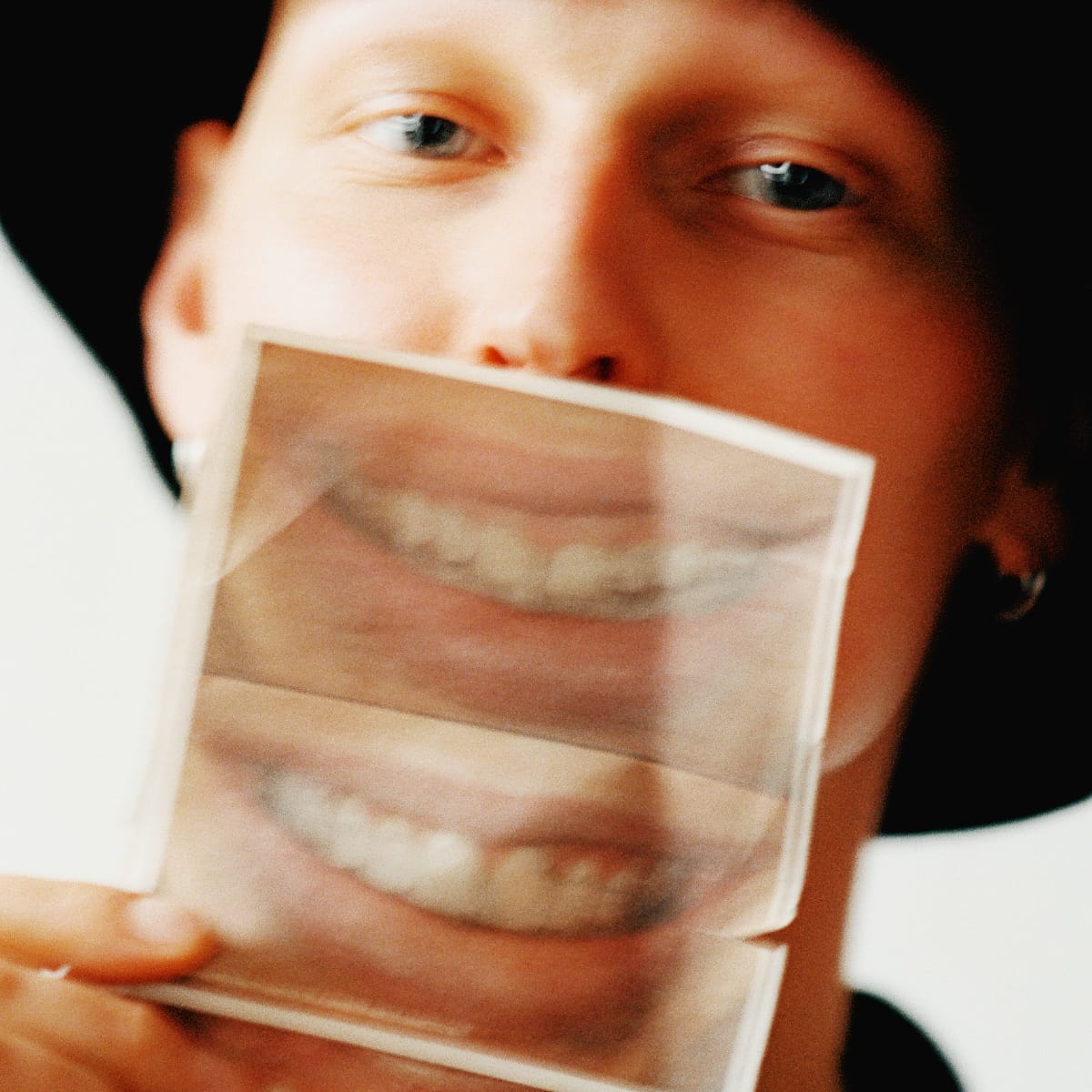 GB holds a strip of photographs of a smiling mouth in front of his own face, eyes visible above it, the pictures slightly out of focus.