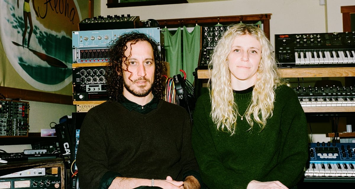 Samur Khouja and Julia Shapiro sit side by side in a studio, racks of synthesizers, keyboards, and outboard gear filling the shelves behind them.