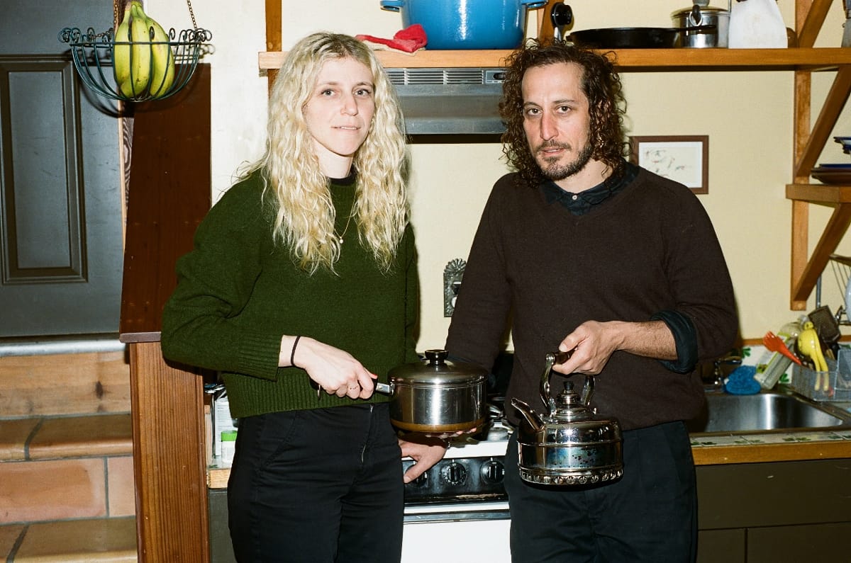 Julia Shapiro and Samur Khouja stand at a kitchen stove holding cookware toward the camera — a lidded steel pot and a chrome kettle — pans and a hanging bunch of bananas behind them.