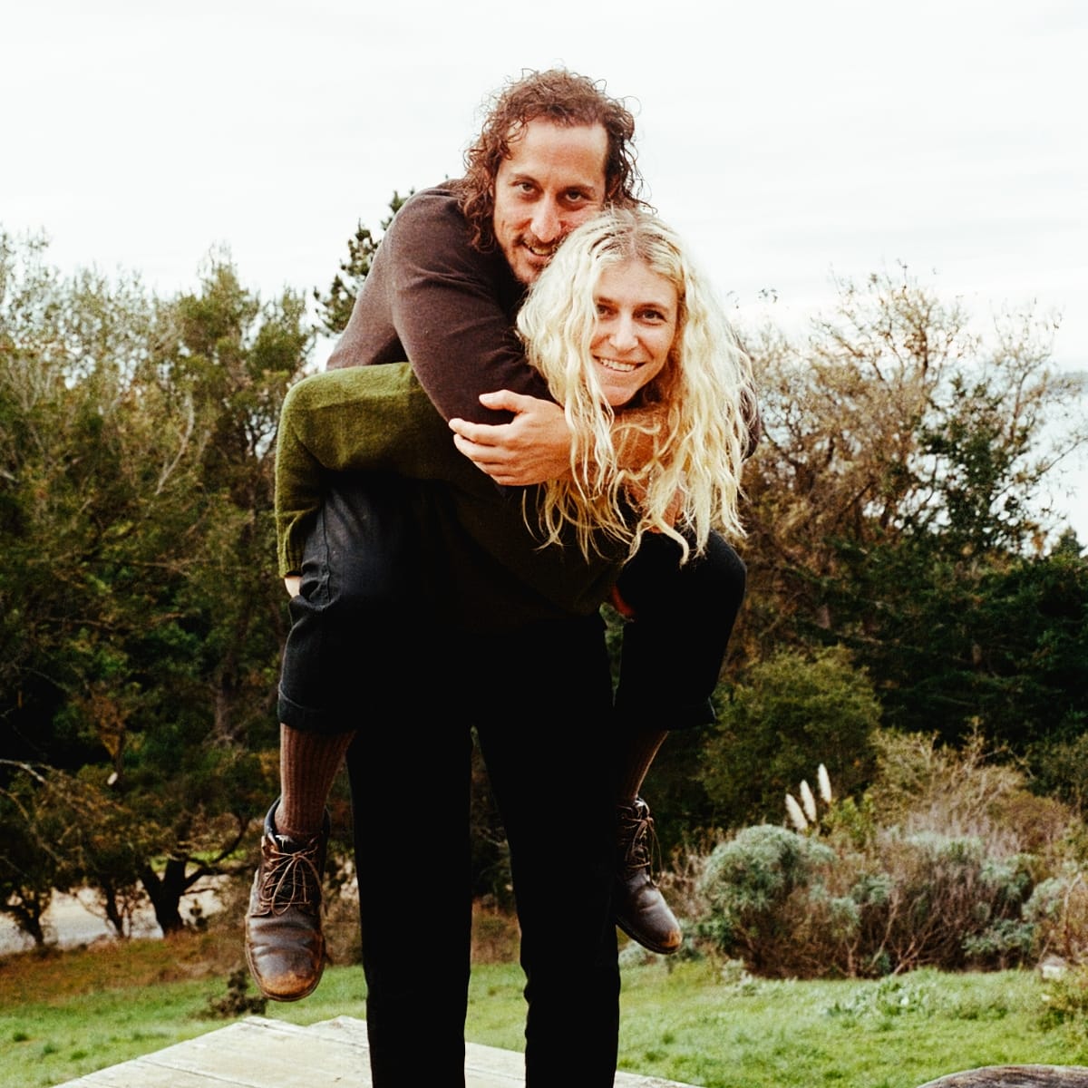 Julia Shapiro carries Samur Khouja piggyback on a wooden deck, both smiling at the camera, with scrubby trees, grass, and a pale sky behind them.